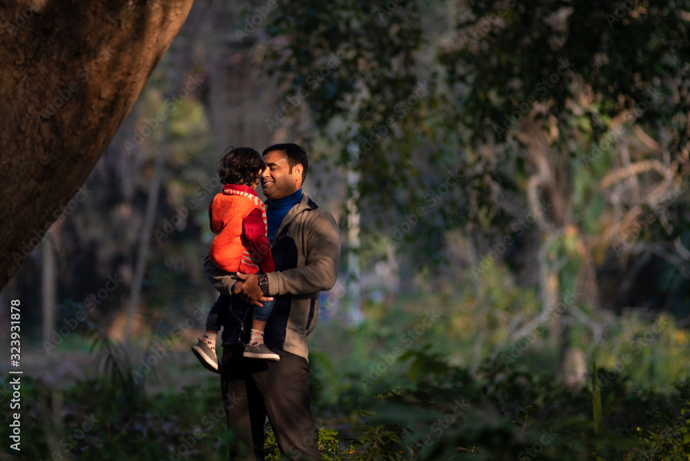 An Indian brunette father and his baby boy in winter garments enjoying themselves in winter afternoon in light and shadow in green forest background. Indian lifestyle and parenthood.