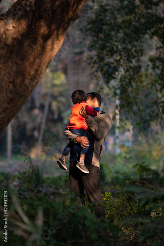 An Indian brunette father and his baby boy in winter garments enjoying themselves in winter afternoon in light and shadow in green forest background. Indian lifestyle and parenthood.