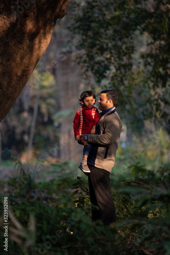 An Indian brunette father and his baby boy in winter garments enjoying themselves in winter afternoon in light and shadow in green forest background. Indian lifestyle and parenthood. © abir