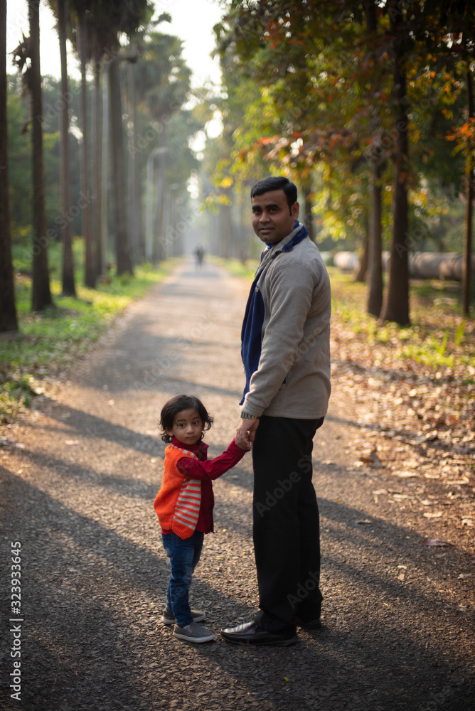Fototapeta premium An Indian brunette father and his baby boy in winter garments standing on a forest path in winter afternoon in natural green background. Indian lifestyle and parenthood.