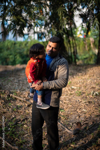 An Indian brunette father and his baby boy in winter garments enjoying themselves in winter afternoon on a  dry grass field in forest background. Indian lifestyle and parenthood. © abir