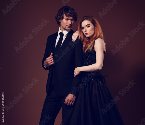 Sexy bright redSexy redheaded hairstyle female woman in fashion black long skirt dress hugging her handsome man in black suit clothing on orange background. Closeup. Toned color portrait