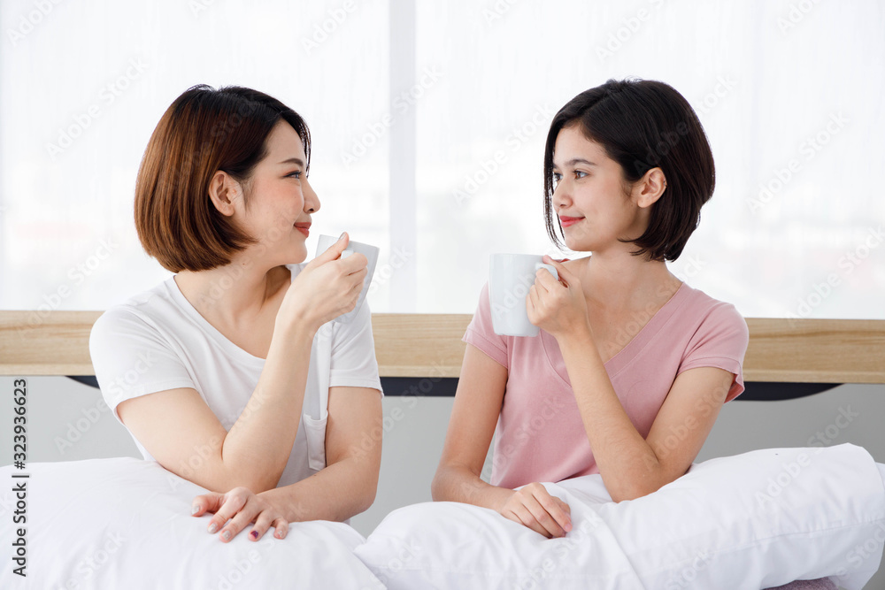 Two Asian women drink beverage from white cup in bedroom. Concept  for teenage or friends activity, lifestyle at home. 