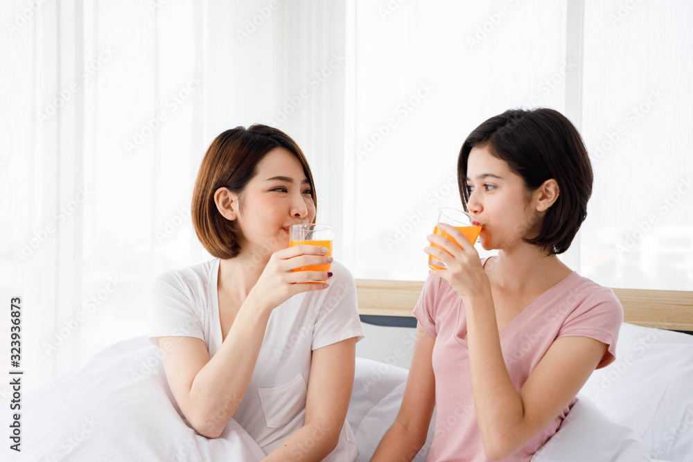 Two Asian women drink glass of orange juice in bedroom. Concept for teenage or friends activity, lifestyle at home with copy space on the right side. Two Asian women drink glass of orange juice in bedroom. Concept for teenage or friends activity, lifestyle at home with copy space on the right side.