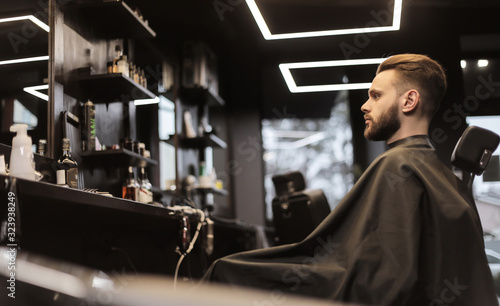 Looking in the mirror. Side photo of a young bearded man looking in the mirror while sitting a soft leather chair in a barbershop.