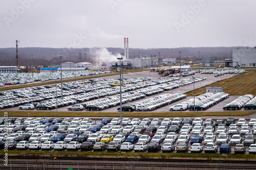 New cars parked in a distribution center on a cloudy rainy day in the autumn and a car factory buildings. Parking in the open air.