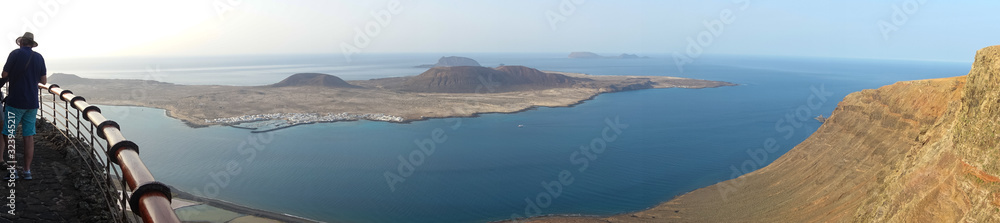 A panoramic view of the volcanic island of La Graciosa meaning graceful in Spanish off Lanzarote,Spain.A male tourist wearing a hat stands by a railing on a cliff edge admiring the scenery.Image