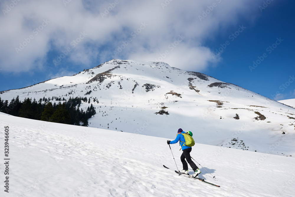 Male hiker, tourist in colorful clothing with backpack hiking on skis up snowy hill on background of bright blue sky and beautiful mountain on frosty sunny day. Winter active lifestyle concept.