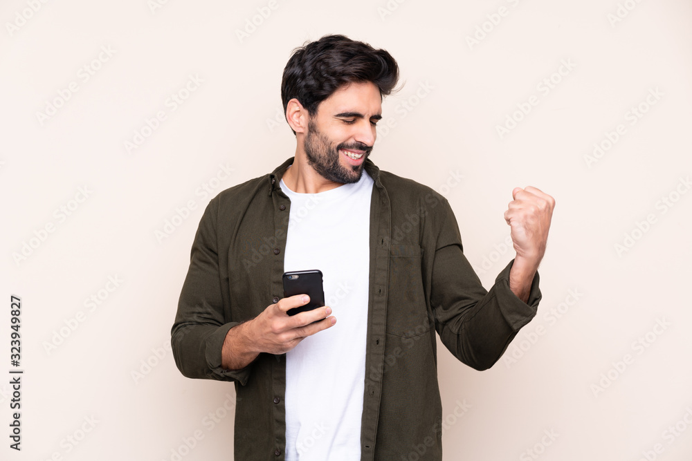 Young handsome man with beard over isolated background with phone in victory position Young handsome man with beard over isolated background with phone in victory position