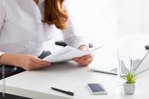 Cropped view of businesswoman holding papers with dossier at table with laptop and smartphone photo