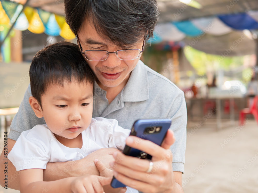 Fototapeta premium Asian little child boy watching smart phone with dad with happy face. Kid and father looking to mobile phone together outdoor.