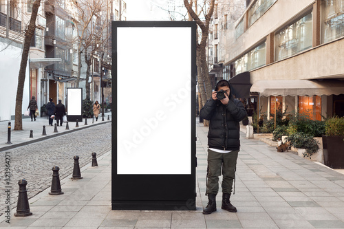 Young photographer stands in front of an empty billboard while taking pictures on the street