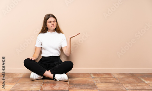Ukrainian teenager girl sitting on the floor holding copyspace with doubts