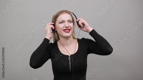 Young blonde girl in black jacket on grey background annoyed woman with headphones listening to music