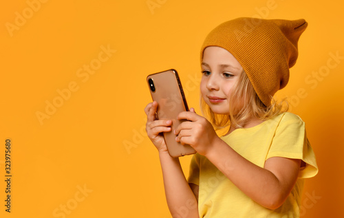 Little blonde kid dressed in t-shirt and hat, posing with smartphone against yellow background. Technology, children, internet. Close-up shot
