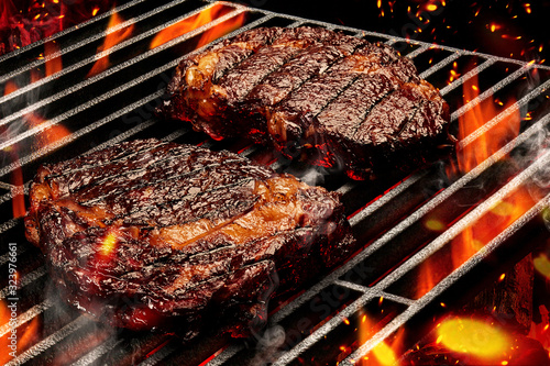 Process of preparing two pork or beef steaks. Meat roasted on metal portable barbecue BBQ grill with bright flaming fire and ember charcoal. Close up