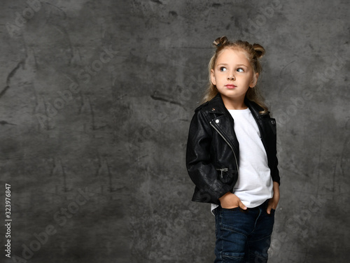 Little cute smiling blond girl in stylish rock style black leather jacket and blue jeans standing over grey concrete background photo