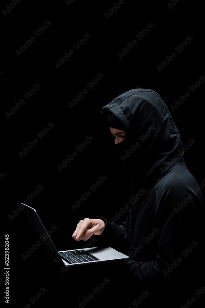 hacker in hood and mask pointing with finger at laptop isolated on black hacker in hood and mask pointing with finger at laptop isolated on black