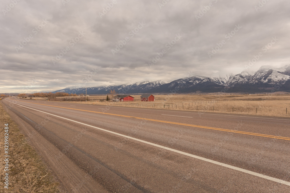 Red house and barn on side of empty highway with open pasture and snow capped mountain range