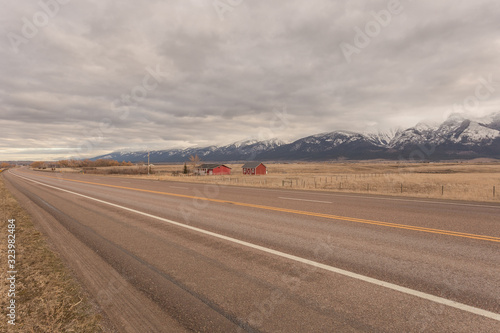 Red house and barn on side of empty highway with open pasture and snow capped mountain range