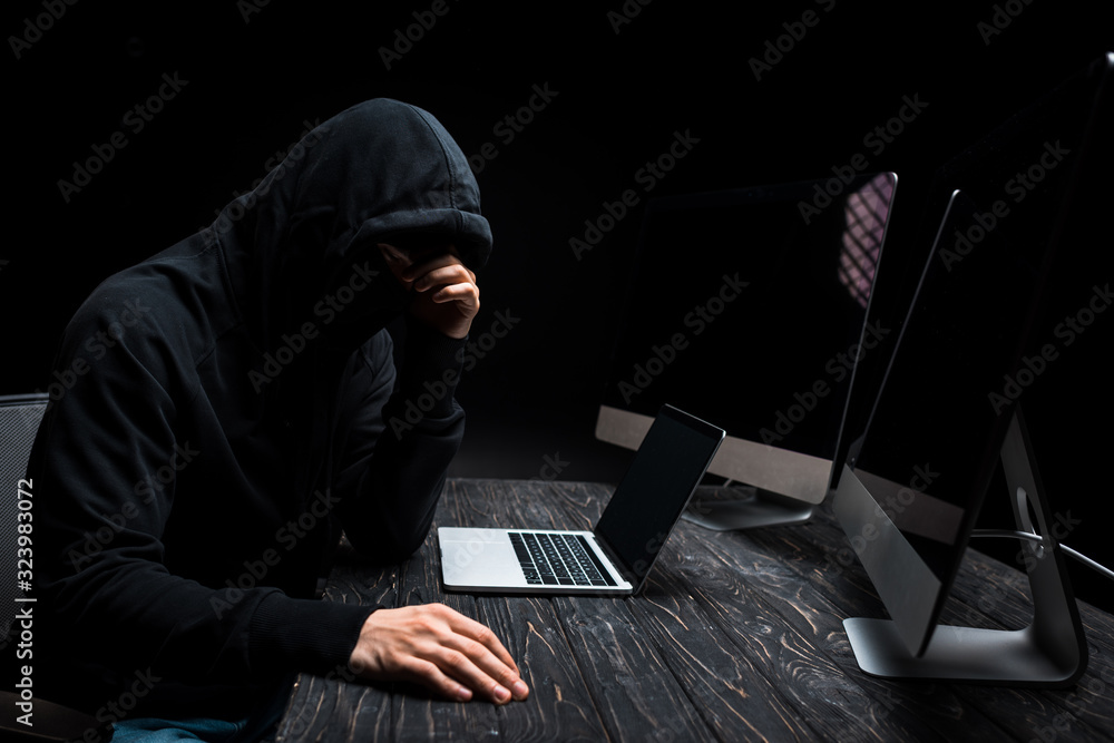upset hacker sitting near laptop and computer monitors with blank screen isolated on black upset hacker sitting near laptop and computer monitors with blank screen isolated on black