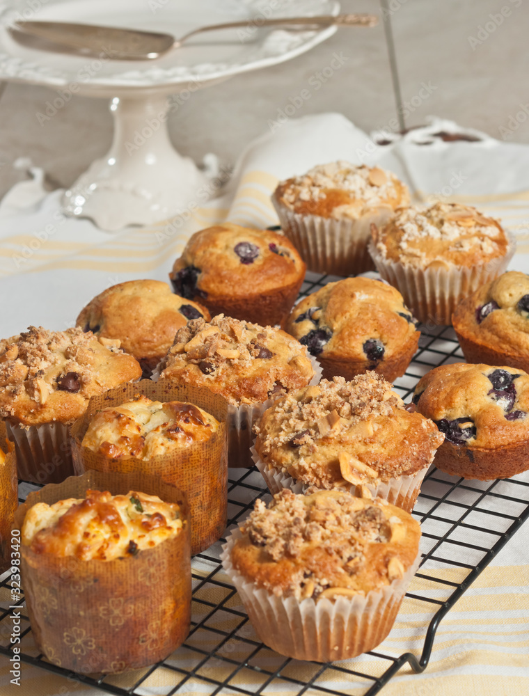 Top, front view of three rows of, variety of freshly baked, homemade muffins, cooling on a wire rack on yellow, white towel and pedestal display dish
