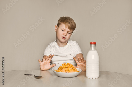 The boy sits at the table and doesn 't want to eat breakfast. Tired of useful food. Moves away from himself a plate of flakes and milk. photo