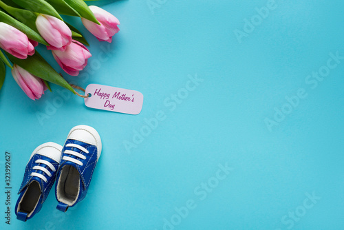 Top view of tulips with happy mothers day lettering on paper label near baby booties on blue background