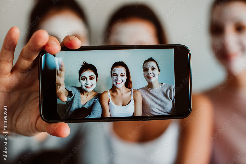 Group of friends applying a revitalizing white mask on their faces. Beauty treatment and skin care. Seated on the sofa taking pictures with a smpartphone together. Close up.