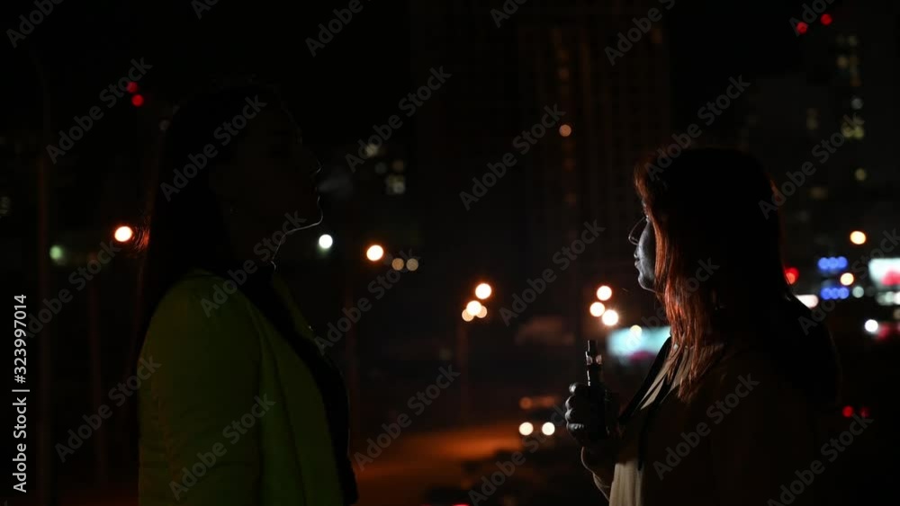 Two girlfriends smoke on the background of the night city. Women smoke cigarettes and vape late at night. Bad habit. Two girlfriends smoke on the background of the night city. Women smoke cigarettes and vape late at night. Bad habit.