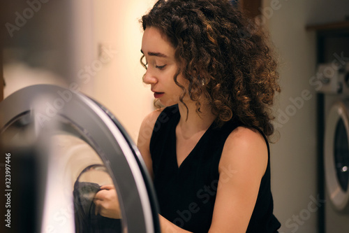Slika na platnu Portrait of attractive brunette girl thoughtfully loading clothes into washing m