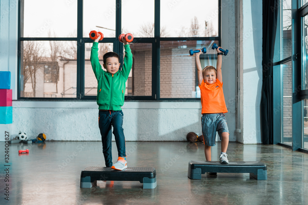 Obraz premium Front view of multiethnic children raising hands with dumbbells while doing Step Aerobics in gym