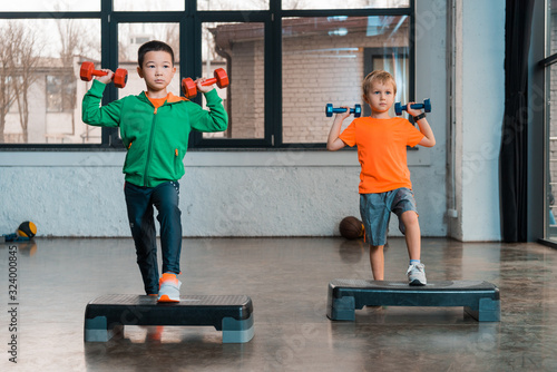 Fototapeta Naklejka Na Ścianę i Meble -  Front view of multicultural children holding dumbbells and doing Step Aerobics in gym