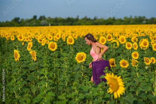 A beautiful brunette in a pink monokini stands in a field of sunflowers. woman in seductive swimsuit walks through the countryside with a luggage bag.