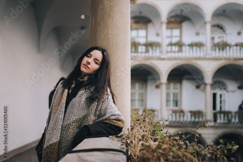 Canvas Print A young black-haired girl in a dark autumn coat and a wide scarf-wrap stands on the balcony with a colonnade and looks looking at the viewer