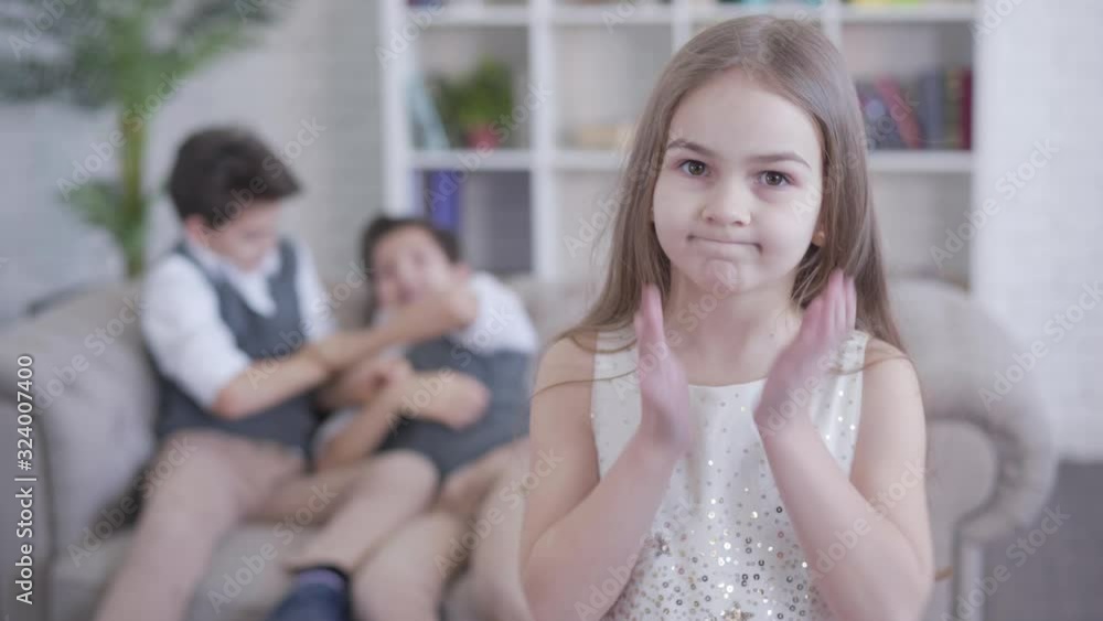 Portrait of pretty little Caucasian girl turning to camera and shaking head as blurred boys fighting at the background. Brunette child with brown eyes shocked with behavior of her twin brothers. Portrait of pretty little Caucasian girl turning to camera and shaking head as blurred boys fighting at the background. Brunette child with brown eyes shocked with behavior of her twin brothers.