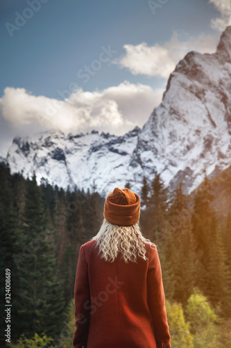 Attractive blonde girl in a red knitted hat stands with her back to the camera on a background of mountains. Travel and tourism concept.