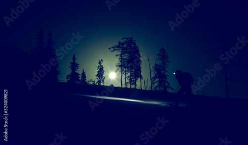 Winter mountain tourism. Night climbing of a tourist on snowshoes with a lamp on the head and a backpack. Moonlight illuminates the road. Light noise and dark night.