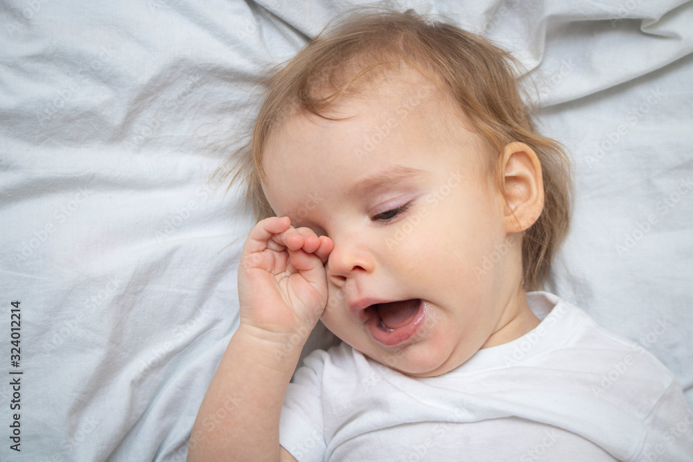 One year old baby girl boy wants to sleep, rubs his eyes with his hand and yawns on white sheets from above. Sleep training mode. One year old baby girl boy wants to sleep, rubs his eyes with his hand and yawns on white sheets from above. Sleep training mode.