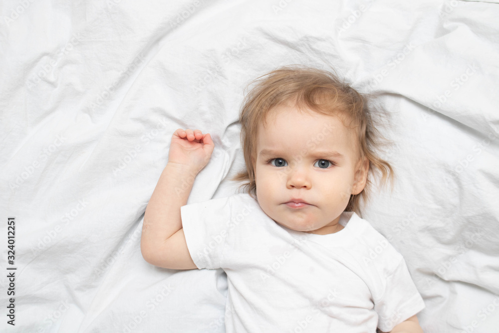 the baby toddler lies on the white sheets of the bed and looks at the camera seriously. Portrait girl one year old boy. the baby toddler lies on the white sheets of the bed and looks at the camera seriously. Portrait girl one year old boy.