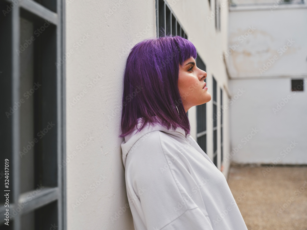 Side view of pensive fashion woman with purple hairstyle leaning on white wall and thoughtfully looking away in bright day Side view of pensive fashion woman with purple hairstyle leaning on white wall and thoughtfully looking away in bright day