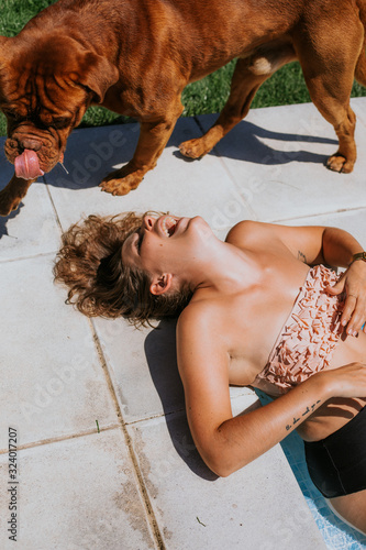 From above of joyful tanned woman in swimsuit laughing while lying on floor and playing with big brown dog on sunny day 