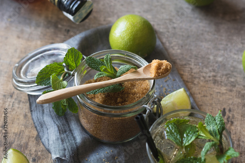 Overhead brown sugar in a jar near fresh limes and peppermint leaves placed on napkin on a wooden table