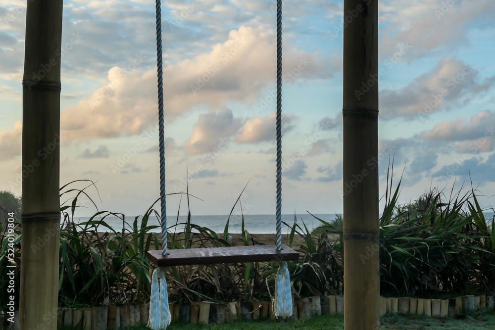 Fototapeta premium A wooden swing with the view on Nyang Nyang Beach. The swing has very simple construction. There is green grass around the swing. Hidden getaway. Collecting happy moments.