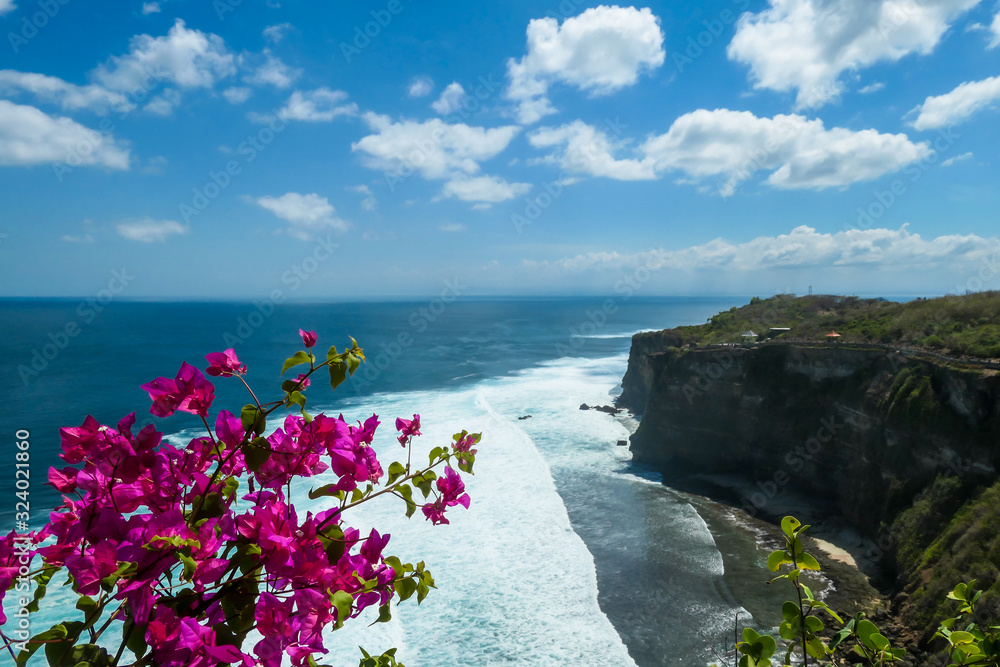 Purple flowers overgrowing the cliff with a view on a small Uluwatu temple, Bali, Indonesia.  The waves are rushing to the shore, making the water bubbly. Steep and dangerous fall. Hidden gem