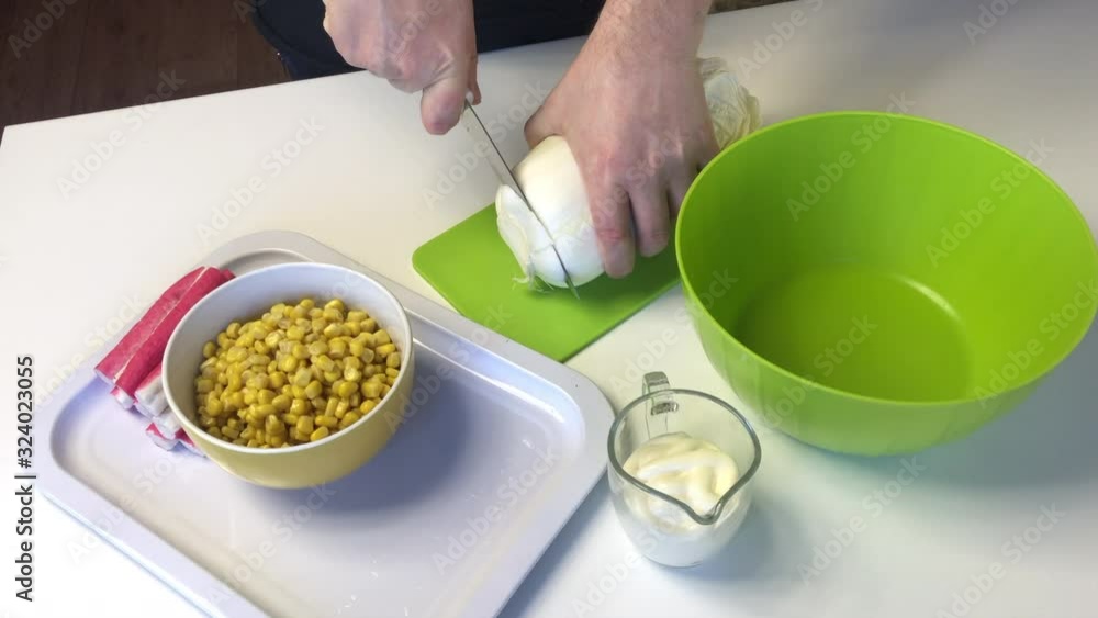 A man makes a salad of Beijing cabbage. Slices cabbage with a knife on a cutting board. Next to the table are other ingredients. Corn, crab sticks and mayonnaise. A man makes a salad of Beijing cabbage. Slices cabbage with a knife on a cutting board. Next to the table are other ingredients. Corn, crab sticks and mayonnaise.