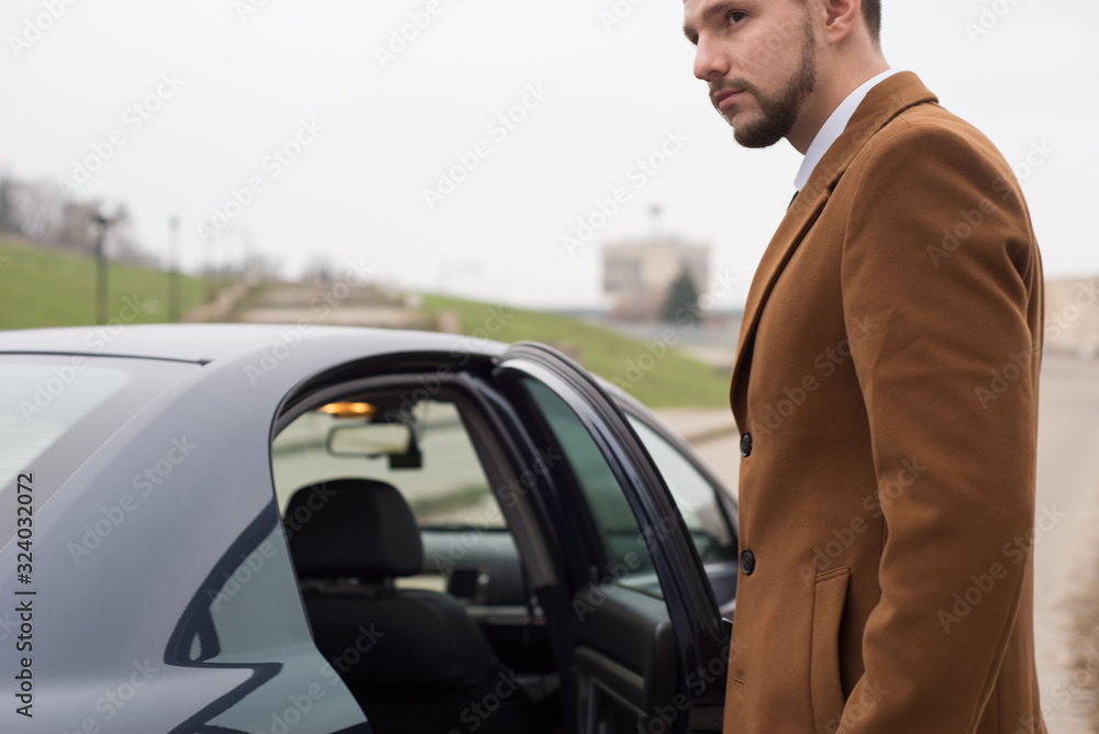 Portrait of a young guy in business clothes, a businessman of thirty years opens the car door to exit. In an interesting authoring
