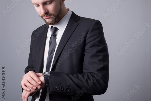 Portrait of a bearded guy, twenty-five years old, in a business suit, looks at his watch. Simple minimalistic business concept. On a gray background