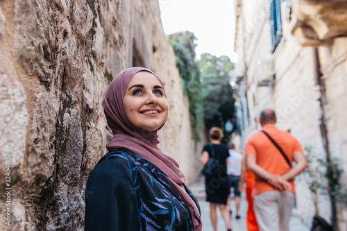 Portrait of beautiful woman with hijab. She is happy and relaxed, while tourist passing by her in narrow old street of Dubrovnik. 2018.