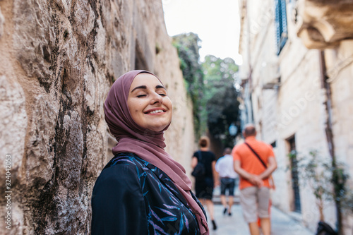 Portrait of beautiful woman with hijab. She is happy and relaxed, while tourist passing by her in narrow old street of Dubrovnik. 2018.